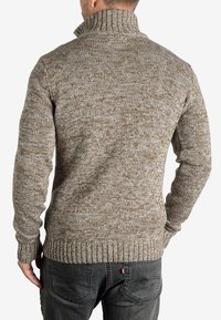Gestrickter Pullover in Braun und Creme, mit hohem Kragen, strukturiertem Muster und gerippten Bündchen und Saum. Getragen mit dunklen Jeans.