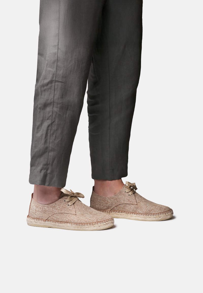 Piernas con pantalones cortos gris oscuro combinados con zapatos casuales beige tejidos con cordones, de pie sobre un fondo claro y simple.