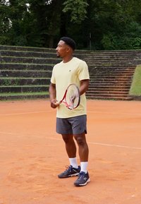 Tennisspiller i en lys gul skjorte og grå shorts, som holder et rødt og hvitt tennisslag på en leirbane med grønne trær i bakgrunnen.