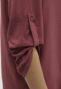 Haut cramoisi texturé avec des manches roulées maintenues par des boutons. Tissu doux avec une apparence légèrement froissée et une coupe décontractée.