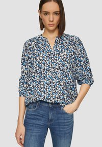 Blouse met een blauw en wit patroon, voorzien van een V-hals, gesmokte details en lange mouwen, gedragen met denim jeans.