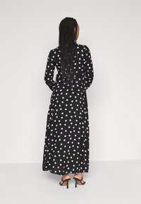 Robe noire longue avec des pois blancs, manches longues et une coupe fluide, portée avec des sandales à talons noires. La vue de dos montre des cheveux tressés.