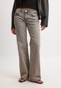 Gaiši pelēki platajām kājām džinsi ar teksturētu apdari, ar pieciem kabatām un metāla pogu aizdari, valkāti ar melnām sandālēm.