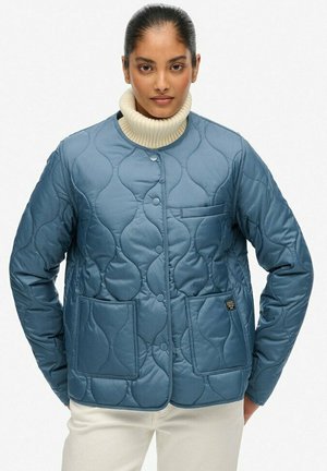 Kvinde iført en blå quiltet jakke med frontlommer og trykknapper over en hvid rullekravesweater.