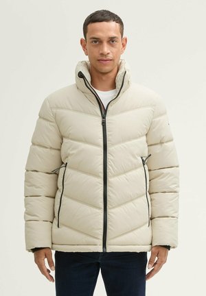 Chaqueta de plumas beige con patrón de flechas, cierre con cremallera y bolsillos laterales. Presenta un cuello alto y cremalleras en contraste de color negro.