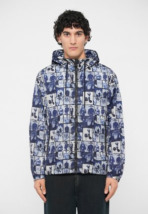 Nuori mies, joka on pukeutunut sinivalkoiseen graafiseen vetoketjulliseen huppariin, jossa on erilaisia valokuvakollaaseja, seisoo tasaisen taustan edessä.