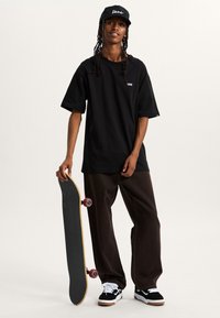 Schwarzes, oversized T-Shirt mit Vans-Logo, lässige braune Hose, schwarz-weiße Sneakers, hält ein Skateboard mit roten Rollen.