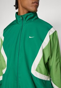 Žalia Nike striukė su baltais ir šviesiai žaliais kampuotais akcentais. Turi aukštą apykaklę ir pilną užtrauktuką, pagaminta iš lygaus, lengvo audinio.