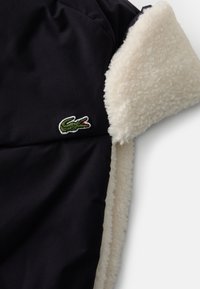Tejido exterior negro con un suave forro de felpa color crema. Presenta un logo bordado de cocodrilo verde en el lateral.