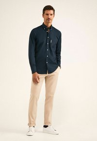 Camicia a maniche lunghe blu navy con colletto a bottoni, abbinata a pantaloni beige e sneakers bianche. Presenta una tasca sul petto e bottoni bianchi.