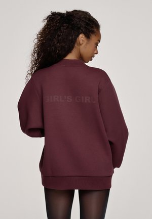 Frau mit lockigem Haar, die ein übergroßes, dunkelmalvenfarbenes Sweatshirt mit dem Aufdruck "GIRL’S GIRL" auf der Rückseite trägt, kombiniert mit schwarzen, durchsichtigen Strumpfhosen.