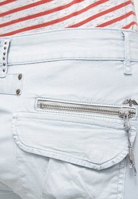 Ljust blå denimbyxor med en dragkedjeförsedd bakficka och dekorativa silvernitar längs midjan. Strukturerad tyg med en slät yta.