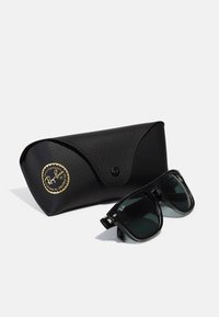 Ochelari de soare pătrati negri cu lentile verzi, având un logo Ray-Ban proeminent, lângă o carcasă texturată neagră cu un emblemă aurie.