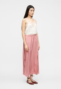 Γυναίκα με μακριά σκουρόχρωμα μαλλιά που φοράει λευκό καλοριφέρ με δαντέλα, ροζ σατέν midi φούστα και καφέ δερμάτινα παπούτσια με κορδόνια, στέκεται μπροστά σε ένα απλό φόντο.