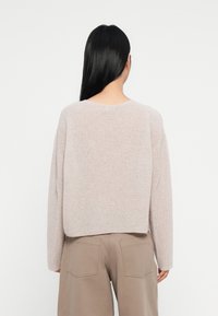 Femeie cu păr lung și negru, îmbrăcată cu un pulover tricotat de un bej deschis și pantaloni taupe, stând cu fața către un fundal alb simplu.