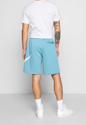 Homme portant un short bleu clair avec un swoosh blanc, un t-shirt blanc, des chaussettes blanches et des baskets blanches, debout dos à la caméra sur un sol gris.