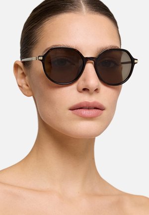 Donna con capelli pettinati all'indietro che indossa occhiali da sole rotondi con montatura tartarugata e lenti scure su uno sfondo semplice.