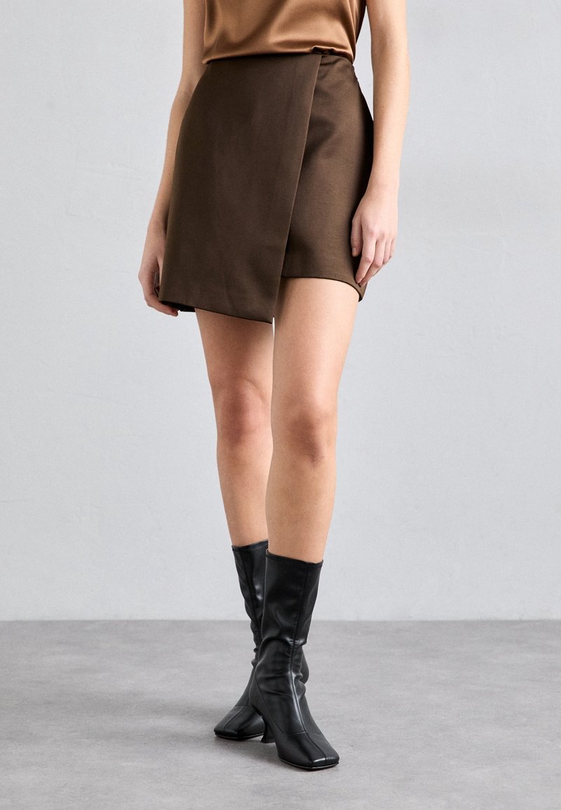 Femme portant une mini-jupe asymétrique marron, un haut marron et des bottes noires à talons mi-mollet, debout sur un sol gris contre un mur clair.