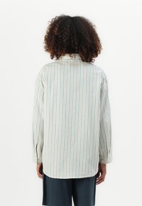 Person med krøllete hår iført en kremfarget langermet skjorte med tynne blå vertikale striper og mørke shorts, som står mot en hvit bakgrunn.