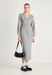 Abito cardigan lungo grigio a maglia con scollo a V, bottoni frontali e texture a coste. Abbinato a una borsa nera e scarpe nere voluminose.