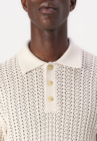 Polo de punto color crema con patrones verticales, un cuello texturizado y tres cierres de botón amarillos.