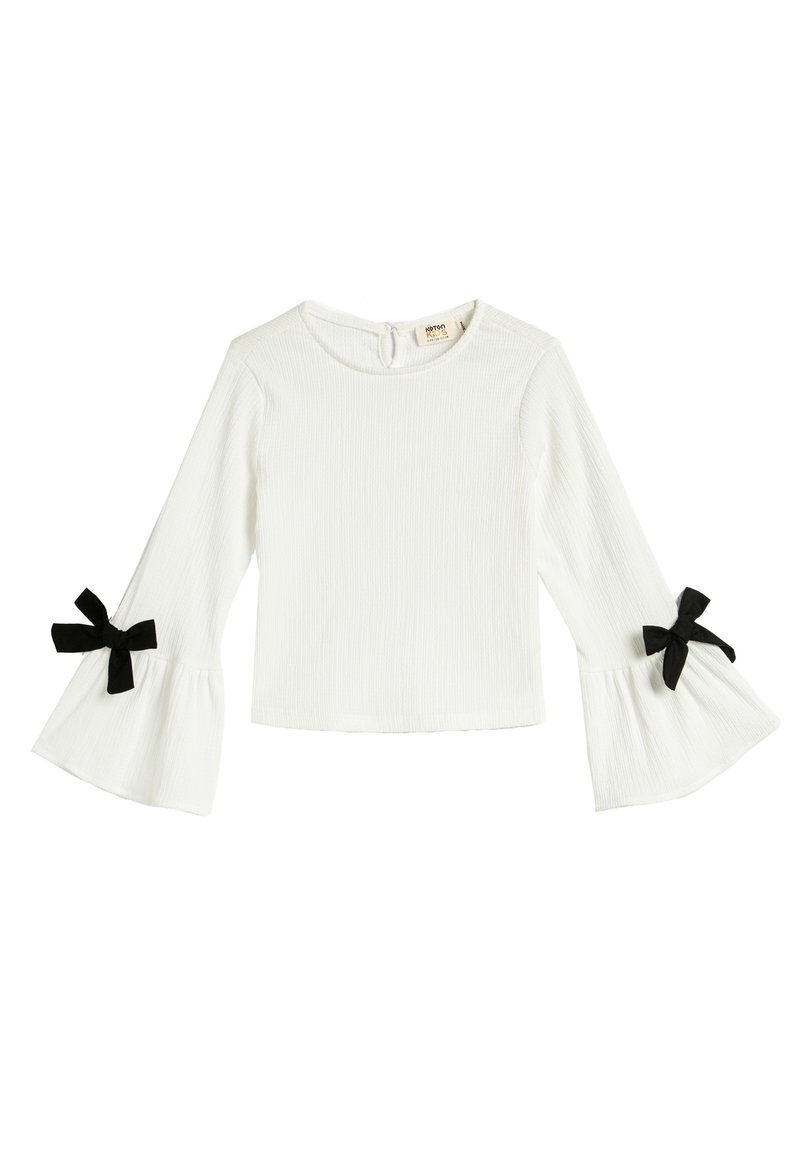 White long-sleeve top with flared sleeves, featuring black bow accents at cuffs and a textured fabric. Round neckline with a back button closure.