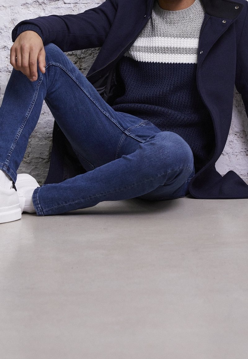 Homme assis par terre portant un jean bleu, des baskets blanches, un pull rayé bleu marine et gris, et un manteau bleu marine foncé devant un mur en briques blanches.