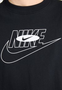 Juodas medvilninis marškinėliai su dideliu baltu Nike logotipu, ryškiu tekstu ir užapvalintu swoosh dizainu, ant vientiso fono.