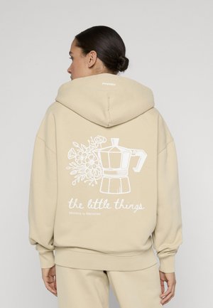 Vrouw draagt een beige hoodie met een illustratie van een koffiepot en bloem en de tekst "the little things Moments to Memories" op de achterkant, kijkt weg.