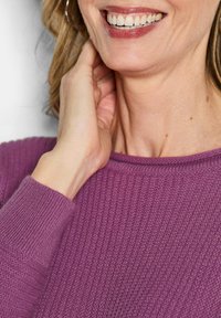 Lächelnde Frau berührt ihren Hals und trägt einen strukturierten lila Pullover.