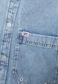 Jasnoniebieska koszula dżinsowa z widocznymi szwami, srebrnymi guzikami i kieszonką z przodu z etykietą "TOMMY JEANS" w kolorze czerwonym i białym.