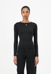 Black long-sleeve top made from stretch fabric, featuring a round neckline and a small white logo on the chest, paired with dark jeans.