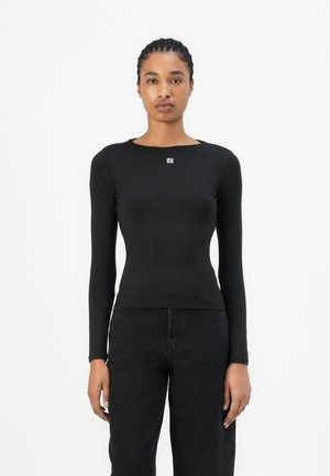 Black long-sleeve top made from stretch fabric, featuring a round neckline and a small white logo on the chest, paired with dark jeans.