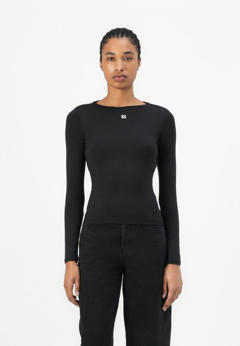 Black long-sleeve top made from stretch fabric, featuring a round neckline and a small white logo on the chest, paired with dark jeans.