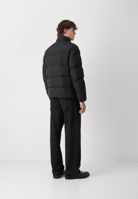 Jachetă puffer neagră cu guler înalt, design matlasat și siluetă scurtă. Asortată cu blugi închiși la culoare și pantofi negri, văzută din spate.