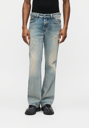 Άτομο που φοράει ξεθωριασμένο μπλε τζιν με φθαρμένα τελειώματα και μαύρα αθλητικά παπούτσια, στέκεται σε λευκό φόντο.