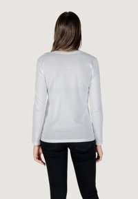 Camicia bianca a maniche lunghe con scollo tondo e silhouette aderente. Indossata con pantaloni neri, che mostrano una texture liscia e leggera.