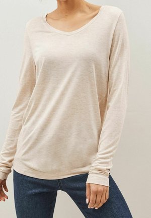 Femme portant un haut à manches longues beige clair à col en V, associé à un jean bleu foncé, debout devant un fond uni.