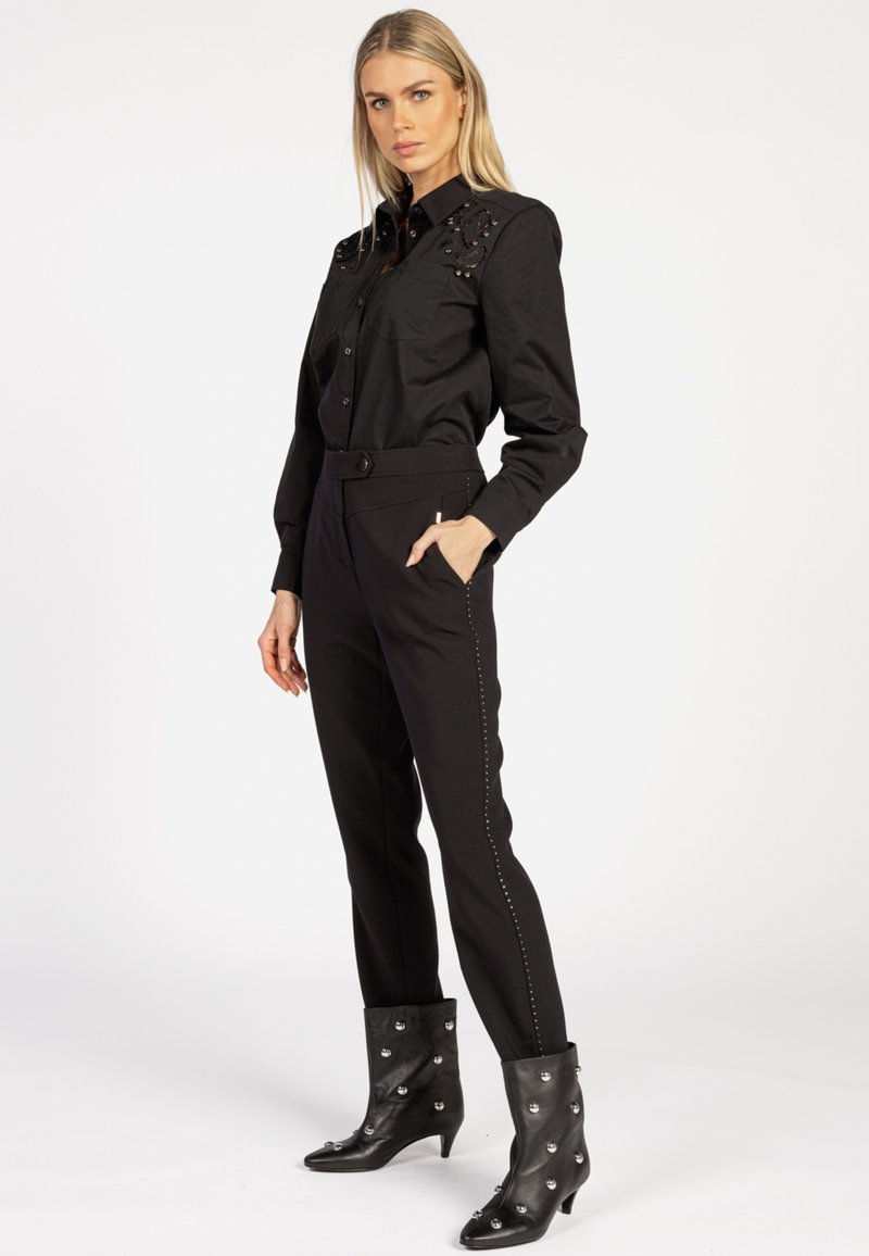 Blonde vrouw die staat, gekleed in een zwarte versierde blouse met knopen, op maat gemaakte broek met zijknopen en zwarte enkellaarsjes met zilveren studs.