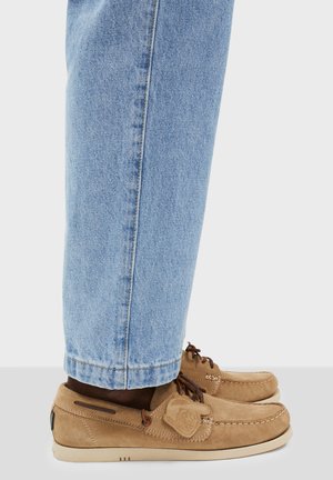 Braune Wildleder-Loafers mit abgerundetem Zehenbereich, braunen Schnürsenkeln und einem dekorativen Logo-Patch. Kombiniert mit hellblauen Denim-Jeans, die am Knöchel leicht umgeschlagen sind.