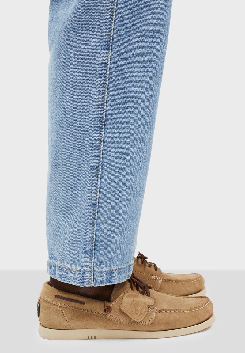 Braune Wildleder-Loafers mit abgerundetem Zehenbereich, braunen Schnürsenkeln und einem dekorativen Logo-Patch. Kombiniert mit hellblauen Denim-Jeans, die am Knöchel leicht umgeschlagen sind.