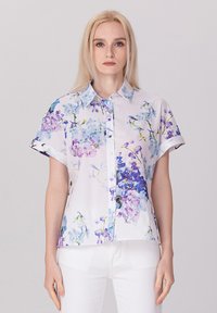 Chemise à manches courtes en tissu léger avec un motif floral bleu et violet sur fond blanc.