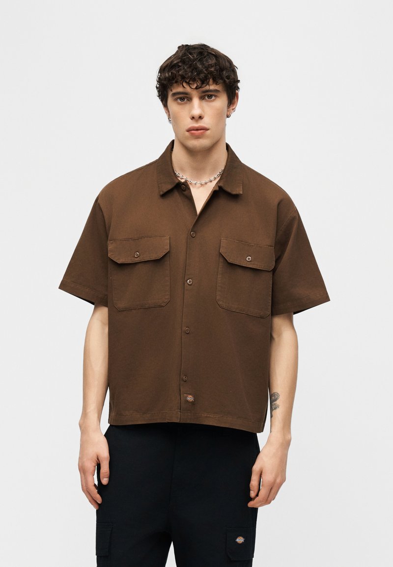 Joven con cabello rizado que lleva una camisa marrón de manga corta con botones y dos bolsillos delanteros, y pantalones negros, contra un fondo claro y liso.