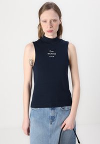 Kleita bez piedurknēm ar augstu apkakli, izgatavota no kokvilnas, tumši zila ar pieguļošu dizainu. Priekšpusē ir izdrukāts balts logo "Tommy HILFIGER" un "Est. 1985".