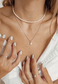 Delikatny naszyjnik z pereł i warstwowy naszyjnik srebrny; widoczny odcień skóry na obojczykach. Jasnozielone paznokcie; pierścionki zdobią palce.