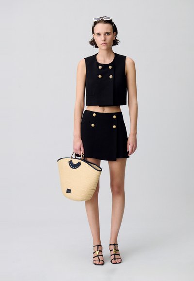 Haut court noir avec des boutons dorés, jupe noire assortie, sac fourre-tout en paille tissée avec des accents noirs, et sandales noires à lanières.