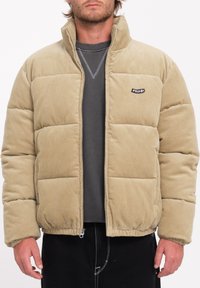 Beige corduroy pufferjack met een hoge kraag, ritssluiting en elastische zoom. Voorzien van een klein merkembleem op de bovenste linkerkant.