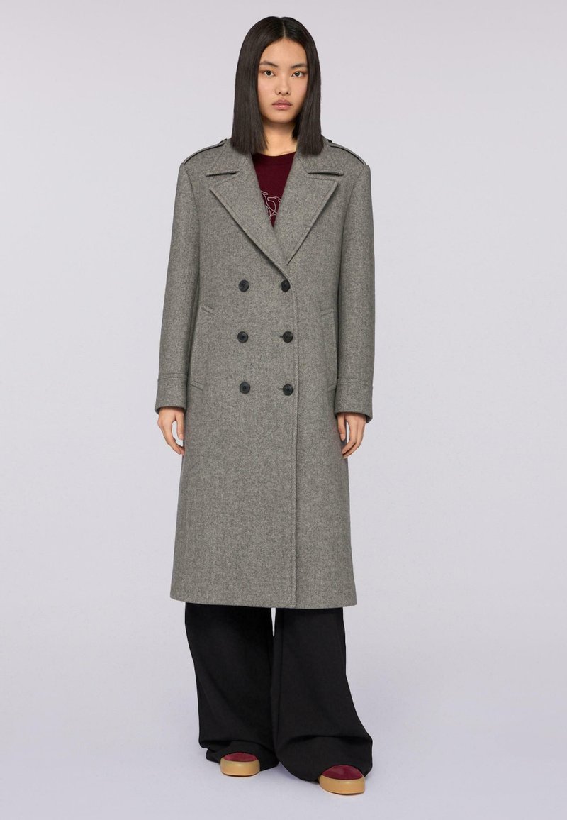 Cappotto grigio di lana doppiopetto con colletto a punta, tasche laterali e bottoni neri, abbinato a pantaloni neri larghi e scarpe con zeppa.