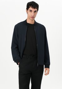 Jachetă de bombardier navy cu fermoar frontal, manșete cu nervuri și o croială ușor relaxată, purtată cu un tricou negru și pantaloni negri cu șnur.