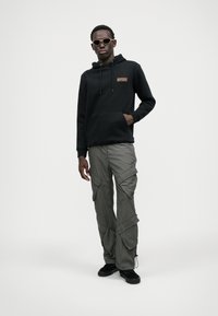 Zwarte hoodie met voorzak en logo, gecombineerd met cargobroeken met meerdere zakken in olijfgroen. Zwarte schoenen en een zonnebril maken de look compleet.